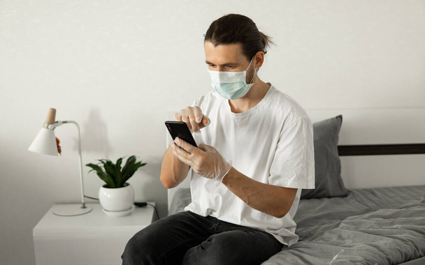 Man in medical mask is using a phone at his home sitting on a bed because of coronavirus epidemic. Remote work during pandemic. Stay home during COVID-19 quarantine concept. - 写真・画像