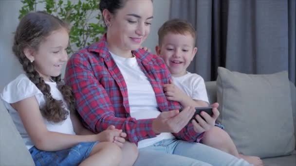 Happy family, mom and cute little kids, mother playing with children at home relaxing use a smartphone cuddling sit on sofa daughter and son laugh, watch funny videos, have fun, enjoy family life moments - Кадры, видео