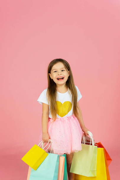 Beautiful cute little girl with long blonde hair, tulle skirt standing in mother`s big shoes with colorful packages isolated on pink background. Expressing true positive emotions to camera. - 写真・画像
