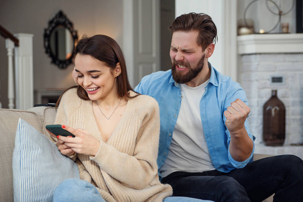 Young wife uses smartphone while sitting on sofa and her husband feels upset behind she. Problems in the family due to addiction to the smartphone. - Fotó, kép
