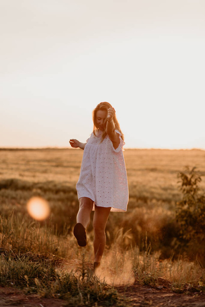 Young beautiful pregnant woman in white dress walking in the wheat orange field on a sunny summer day. Nature in the country. Miracle expectation. Sunset on isolation - Photo, image