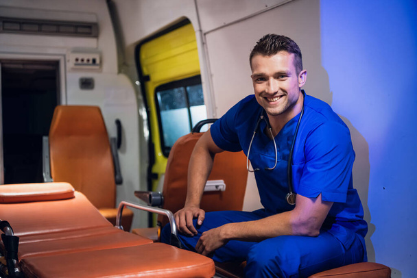 Портрет улыбающегося мужчины в синей медицинской форме, сидящего в машине скорой помощи
 - Фото, изображение