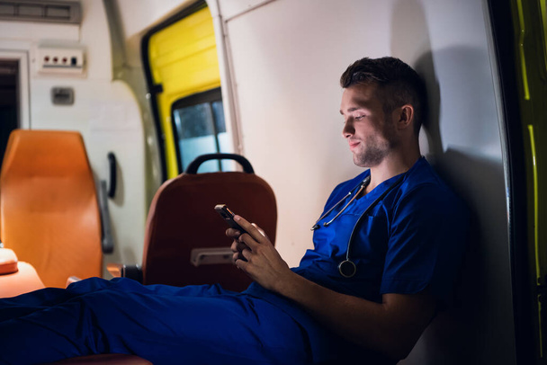 Junger Rettungssanitäter sitzt mit Smartphone im Rettungswagen - Foto, Bild