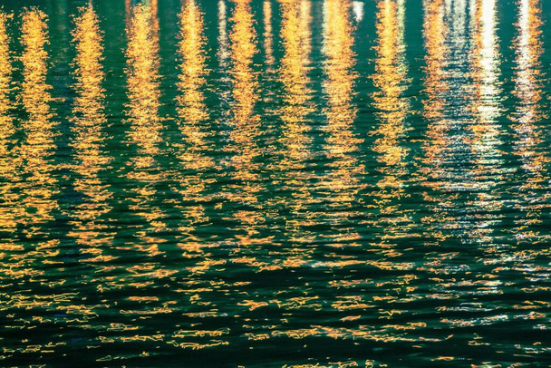 抽象的なテクスチャ、水の暗い表面に黄色の光の反射。ナイトシティ、照明。黄色、オレンジのトーン、垂直ストライプ. - 写真・画像