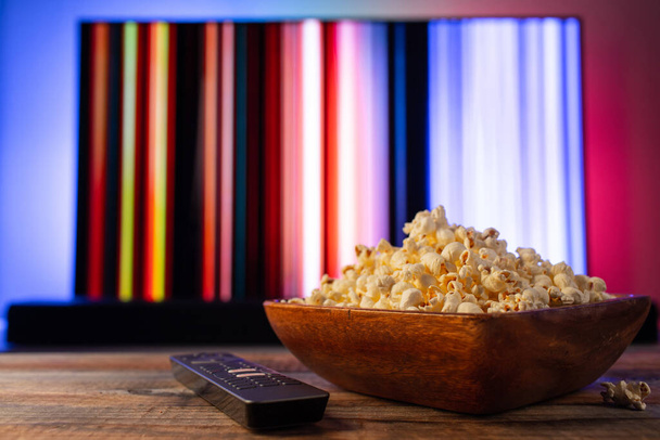 Egy fából készült tál popcorn és távirányító a háttérben a TV működik. Este otthonosan néz egy filmet vagy TV-sorozatot otthon. - Fotó, kép