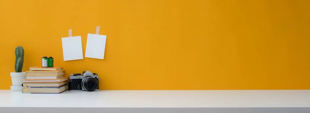 Close up zicht op creatieve werkruimte met camera, boeken, cactus pot en kopieer ruimte op witte tafel met gele wand - Foto, afbeelding