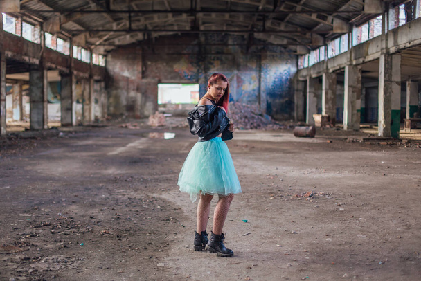 Portrait of a young girl with pink hair dressed in leather jacket and a blue skirt standing inside of collapsed building surrounded by ruins - Fotografie, Obrázek