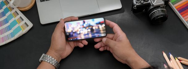 Fotografia aerea del fotografo che guarda video su smartphone orizzontale mentre si siede in uno spazio di lavoro moderno e buio
 - Foto, immagini