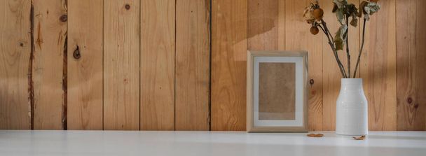 Vista de cerca del moderno diseño interior de la casa rústica con marco de maqueta, jarrón de cerámica y espacio de copia en la mesa blanca con pared de tablón
  - Foto, imagen