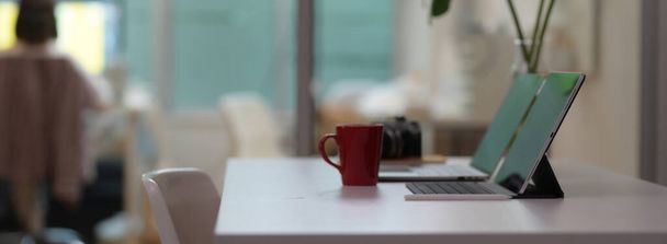 Vista lateral da confortável mesa de escritório com dispositivos digitais, caneca de café, câmera e vaso de planta na mesa branca
    - Foto, Imagem