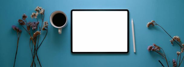 Draufsicht auf kreativen Arbeitsplatz mit leerem Bildschirm-Tablet, Kaffeetasse und trockener Blume auf blauem Schreibtisch    - Foto, Bild