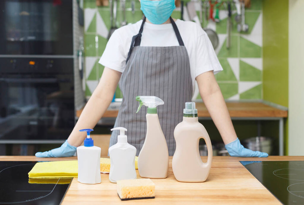 Mujer en máscara y guantes mostrando todos los detergentes ecológicos para la desinfección de la cocina
 - Foto, Imagen