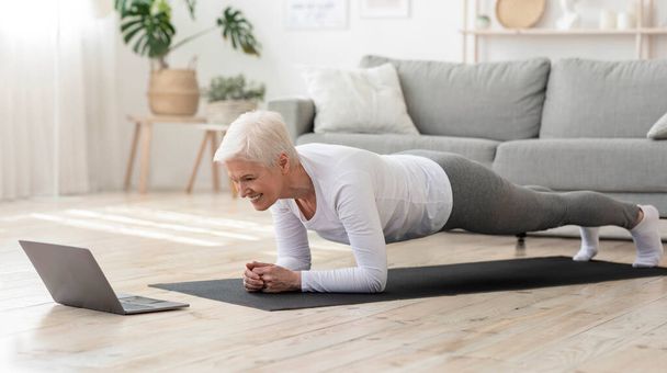 Mujer mayor deportivo haciendo tablón de yoga mientras mira tutorial en el ordenador portátil
 - Foto, Imagen