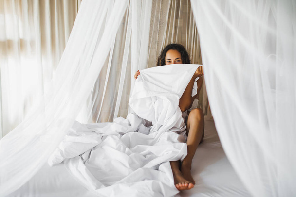 Beautiful tanned young woman awakening in white bed. Happy wake up and start new day. Leisure and rest. Wellbeing and carefree  concept. Long curly hair. Stay home. - Фото, изображение