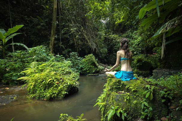 Young Caucasian woman sitting on the rock, practicing yoga and pranayama. Hands in gyan mudra. View from back. Vacation in Asia. Calm and relaxing atmosphere. Tropical nature landscape. Bangli, Bali. - Photo, Image