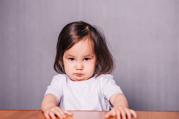 Niña ofendida, niña sentada a la mesa en camiseta blanca sobre fondo gris
. - Foto, Imagen