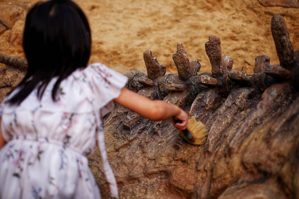 Una ragazza che gioca in una sabbiera con un fossile di dinosauro modellato, spazzolando via la sabbia dal fossile
. - Foto, immagini