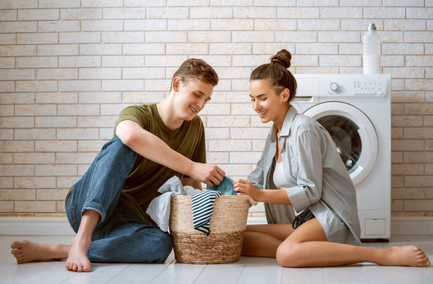 Beautiful young loving couple is smiling while doing laundry at home. - Photo, Image