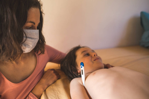 Una mujer con una máscara protectora mide la temperatura del niño, llame a un médico en casa, un niño con fiebre se acuesta en la cama, mamá cuida al bebé durante una enfermedad. - Foto, Imagen