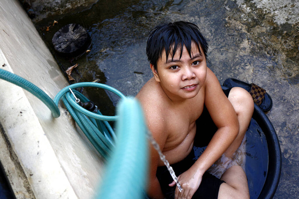 Фото молодого азиатского мальчика, охлаждающегося в водяном бассейне с водяным шлангом, как импровизированный душ, чтобы побороть летнюю жару
. - Фото, изображение