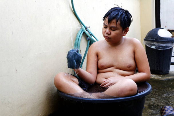 Φωτογραφία ενός νεαρού Ασιάτη που δροσίζεται σε μια λεκάνη νερού με σωλήνα νερού ως πρόχειρο ντους για να νικήσει τη ζέστη του καλοκαιριού. - Φωτογραφία, εικόνα