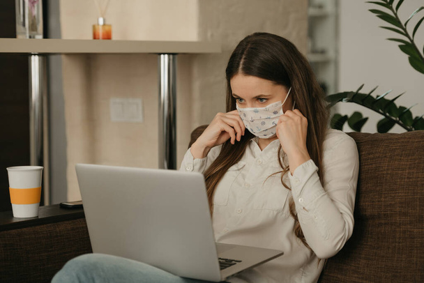 Trabalho remoto. Uma mulher em uma máscara facial médica estudando remotamente em seu laptop durante a quarentena para evitar o coronavírus disseminado. Uma menina colocando uma máscara facial em casa durante a pandemia de COVID-19
 - Foto, Imagem