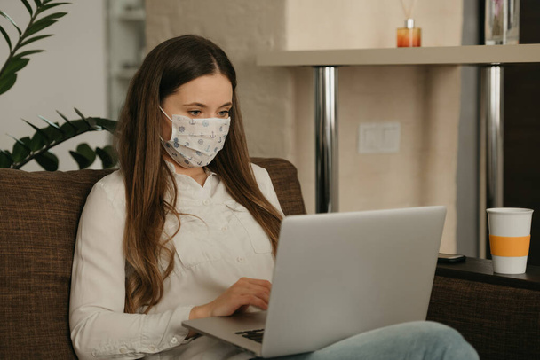 Praca zdalna. Kobieta w masce medycznej pracująca zdalnie na laptopie podczas kwarantanny, by uniknąć rozprzestrzeniania się koronawirusu. Dziewczyna w masce na twarz w domu podczas pandemii COVID-19 - Zdjęcie, obraz
