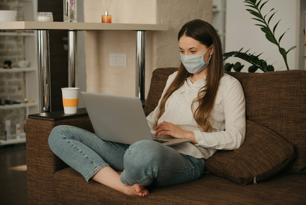 Praca zdalna. Kobieta w masce medycznej pracująca zdalnie na laptopie podczas kwarantanny, by uniknąć rozprzestrzeniania się koronawirusu. Dziewczyna pracująca z domu w masce na twarz podczas pandemii COVID-19 - Zdjęcie, obraz