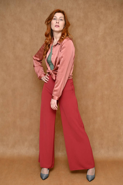 A pretty model is wearing red pants and a pink blouse. Full-length vertical portrait of a young Caucasian red-haired woman on a beige background in various poses. - Fotó, kép
