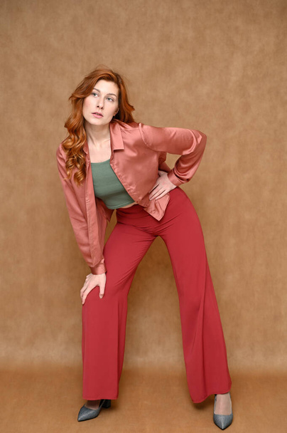 Nice girl model dressed in red pants and a pink blouse. Full-length vertical portrait of a young Caucasian red-haired woman on a beige background in various poses. - Fotografie, Obrázek