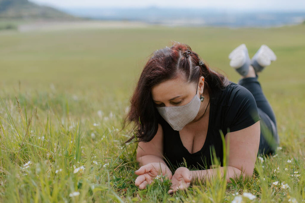 Молода жінка в чорному в захисній медичній масці лежить на зеленій траві. Весняне літо. Дівчина тримає полуничні квіти в руках. Дівчина не дивиться в рамку
. - Фото, зображення