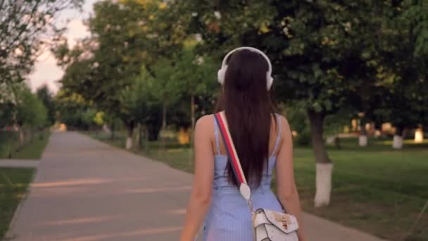 Charming young woman is walking down the street in the park, listening to the music in her earphones. Summertime, outdoors.  - Felvétel, videó