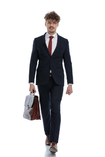 Jistý podnikatel drží svůj kufřík, zatímco na sobě oblek a šlápnutí na bílém pozadí studia - Fotografie, Obrázek