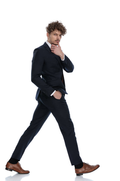 Sivunäkymä huomaavainen liikemies kädestä leuka ja taskussa, kun yllään puku ja kävely valkoinen studio tausta
 - Valokuva, kuva