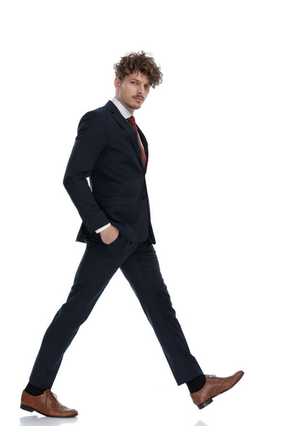 Вид сбоку серьезного бизнесмена, держащего обе руки в карманах во время ношения костюма и прогулки на белом фоне студии
 - Фото, изображение