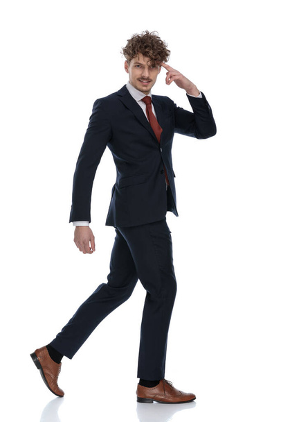 Seitenansicht eines lächelnden Geschäftsmannes, der auf seinen Kopf zeigt, während er Anzug trägt und auf weißem Studiohintergrund läuft - Foto, Bild