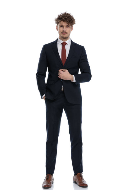 Serious businessman unbuttoning his jacket with hand in pocket while wearing suit and standing on white studio background - Zdjęcie, obraz