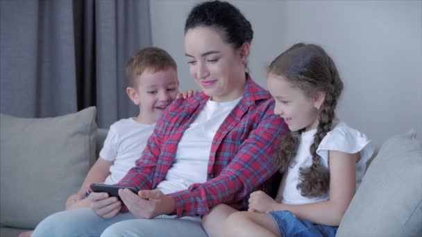 Šťastná rodina, maminka a roztomilé malé děti, matka si hraje s dětmi doma relaxaci pomocí chytrého telefonu mazlení sedět na pohovce dcera a syn se zasmějí, sledovat vtipná videa, bavte se, užijte si rodinné chvíle - Záběry, video