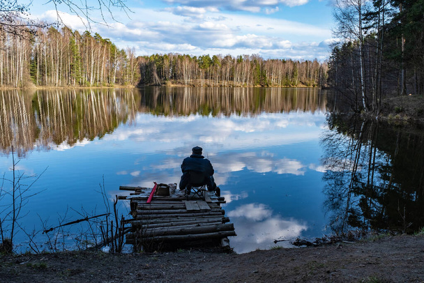 Fisherman on a log pier against the background of the forest and a beautiful reflection of the forest and clouds in the water in early spring.  - Foto, immagini