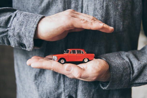 Male hands holding and protecting a red toy car. Conceptual image of insurance and road safety. - Foto, Bild