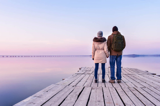 Een man en een vrouw staan met hun rug op de pier, die is gemaakt van grijs hout, en houden elkaars handen vast tijdens zonsondergang, kijkend naar de hemel van roze en lila. De lucht wordt weerspiegeld in een perfect glanzend oppervlak van het water. - Foto, afbeelding