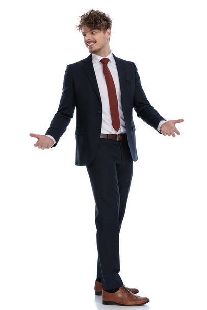 Πλευρική άποψη του θετικού επιχειρηματία αναζητούν πάνω από τον ώμο και εξηγώντας, ενώ φορώντας κοστούμι και στέκεται σε λευκό φόντο στούντιο - Φωτογραφία, εικόνα