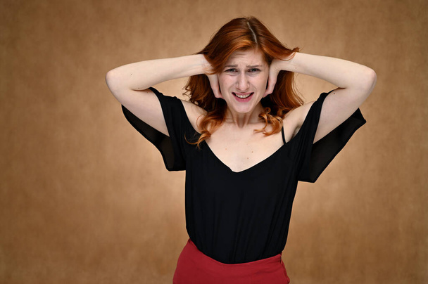 Model actress shows different emotions while talking on camera in the studio. Photo of a pretty caucasian young woman with minimal makeup in a black blouse with long red hair on a beige background. - Foto, Bild