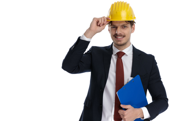 Feliz hombre de negocios sosteniendo portapapeles mientras fija su casco de construcción y el uso de traje, de pie sobre fondo de estudio blanco
 - Foto, Imagen
