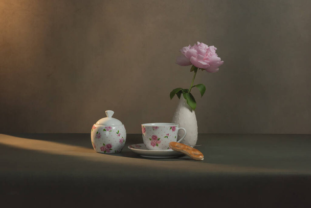 Tazza da tè con piattino e biscotto, zuccheriera e vaso bianco con peonia su un tavolo in una stanza grigia alla luce del sole del mattino
. - Foto, immagini