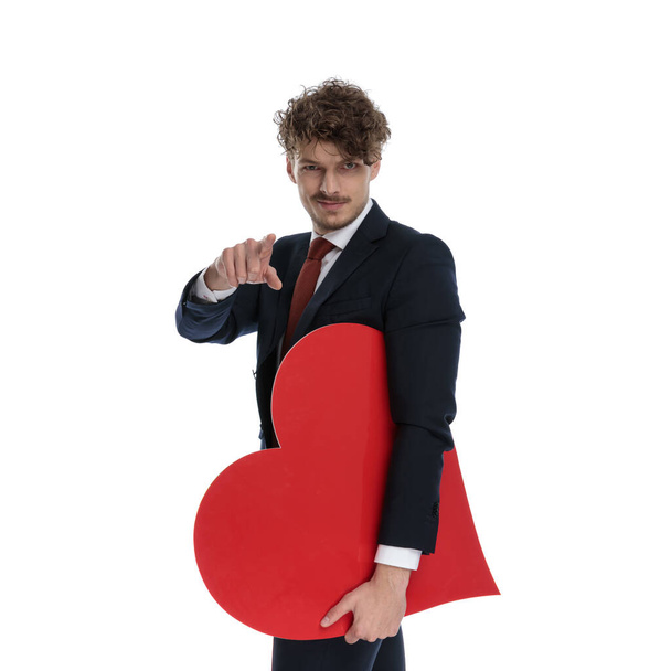 Magabiztos üzletember tartja szív alakú és mutató viselése közben öltöny és álló fehér stúdió háttér - Fotó, kép
