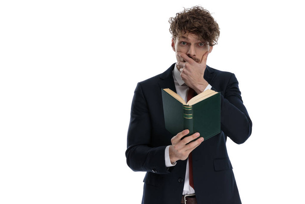 Sorprendido hombre de negocios leyendo libro y cubriéndose la boca mientras llevaba traje y de pie sobre fondo blanco del estudio
 - Foto, Imagen