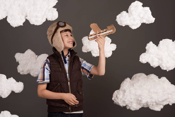een jongen speelt met een kartonnen vliegtuig en droomt ervan piloot te worden, gekleed in een retro jasje en helm met bril, wollen wolken, grijze achtergrond, bruin getint - Foto, afbeelding