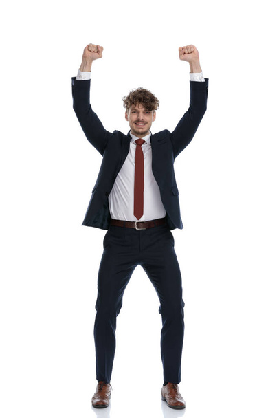 Hombre de negocios alegre sonriendo y celebrando con los dos puños en alto mientras usa traje y de pie sobre fondo blanco del estudio
 - Foto, Imagen