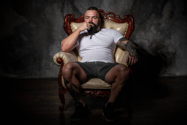 brutal man with beard and tattoos is sitting in a chair. hairstyle shaved whiskey and dreadlocks - Photo, Image